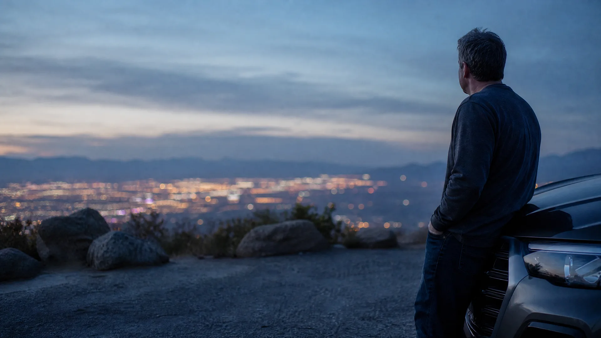 A man in a dark sweater leans against the front of an SUV on a gravel overlook at dusk, facing a distant lit city below with rocks and low shrubs nearby.