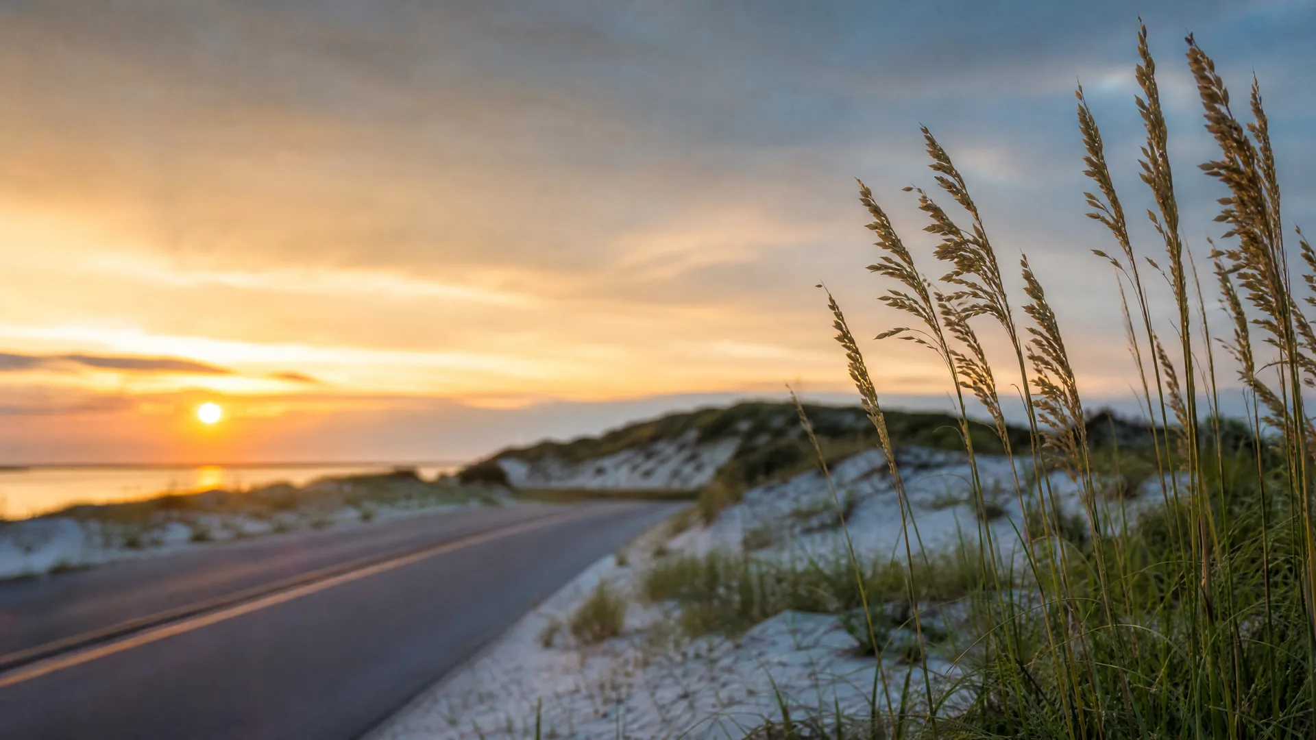 A paved road curves along sand dunes with sea oats and grasses in the foreground as the sun sets low over water in the distance.