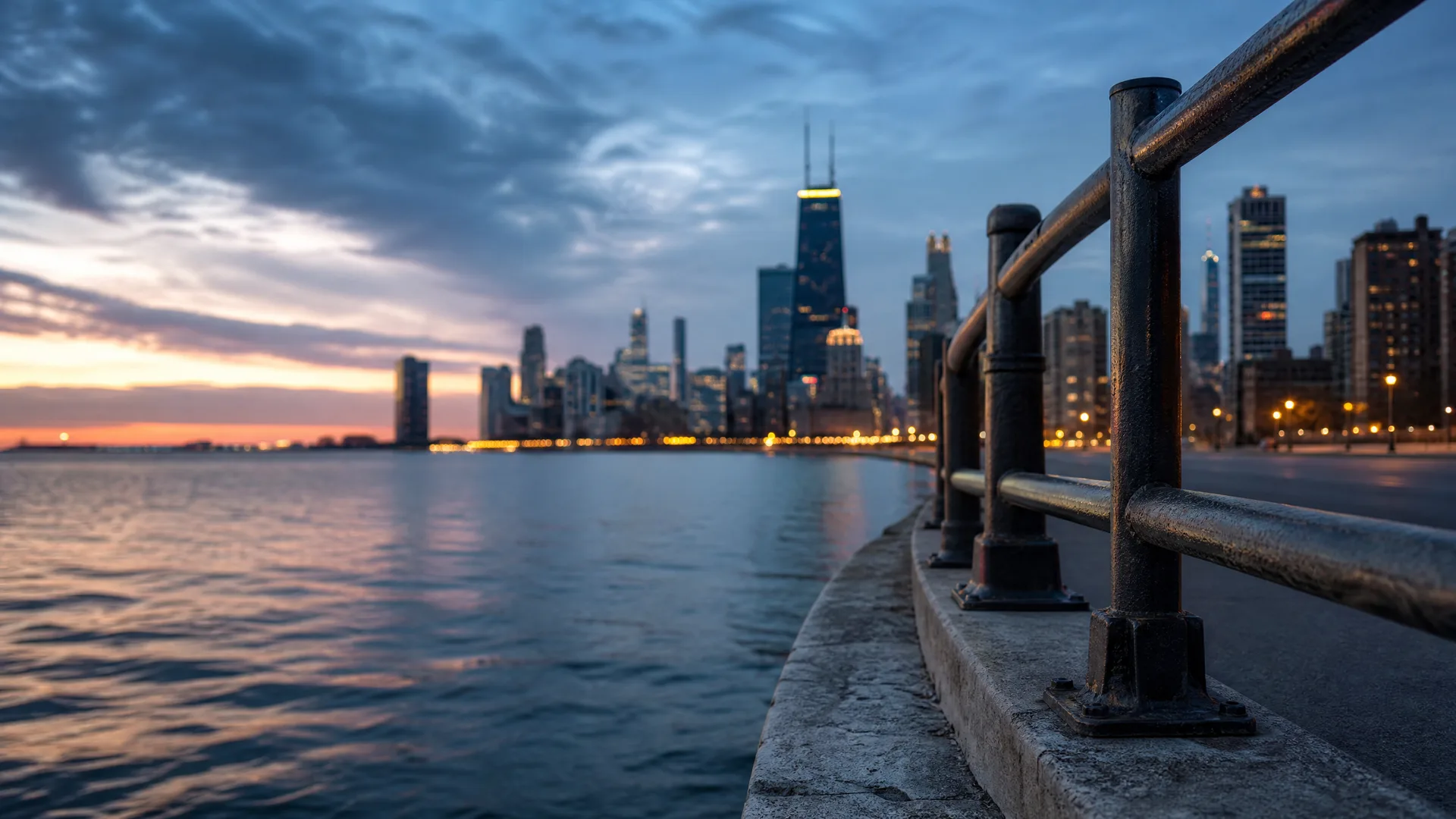 A metal railing on a concrete waterfront ledge in the foreground, with a large body of water at left and a lit city skyline of high-rise buildings under a cloudy twilight sky.