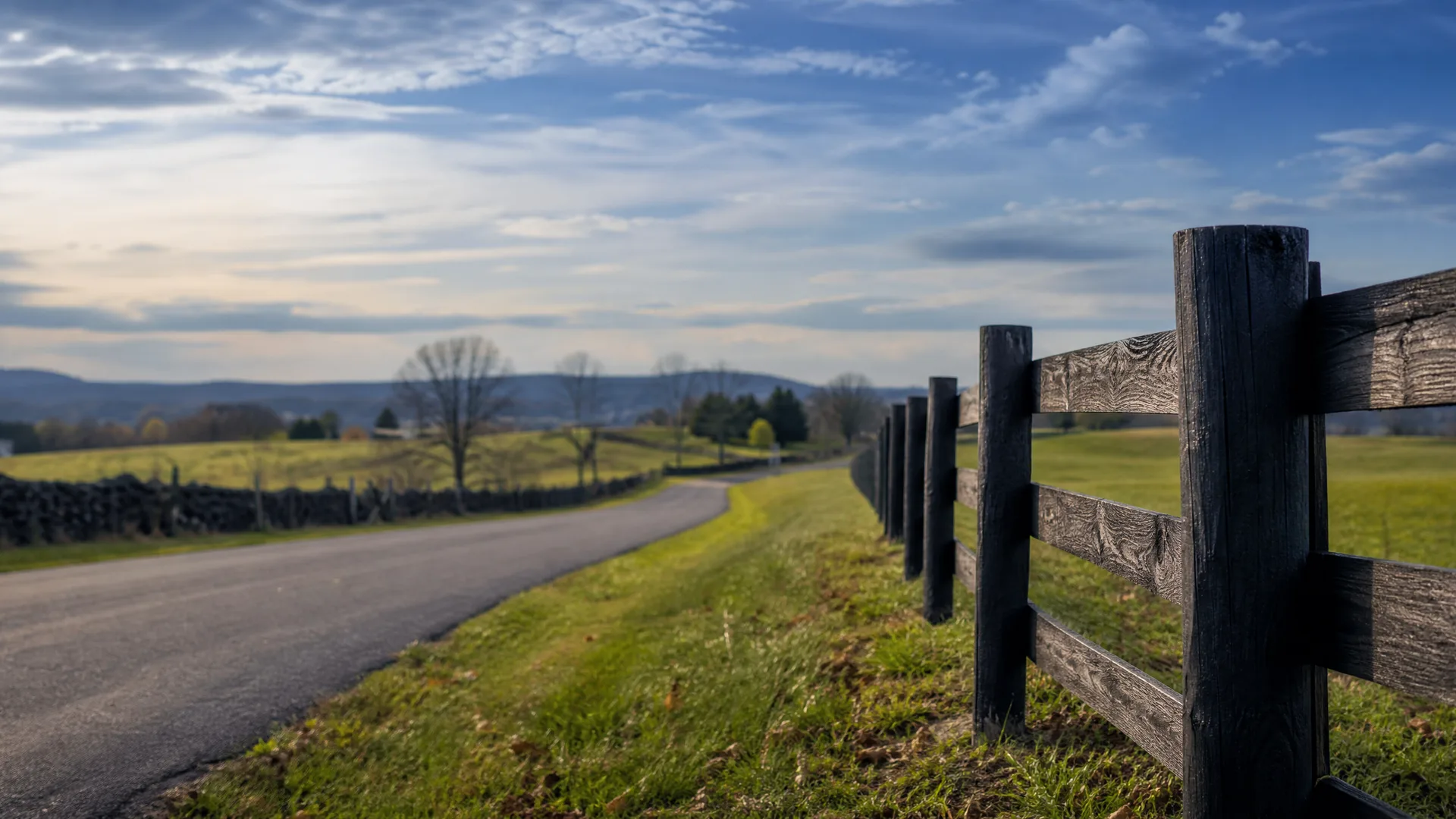 A dark weathered wooden post-and-rail fence runs along the right side of a paved road curving through grassy fields, with bare trees, a low stone wall, and distant hills under a partly cloudy sky.